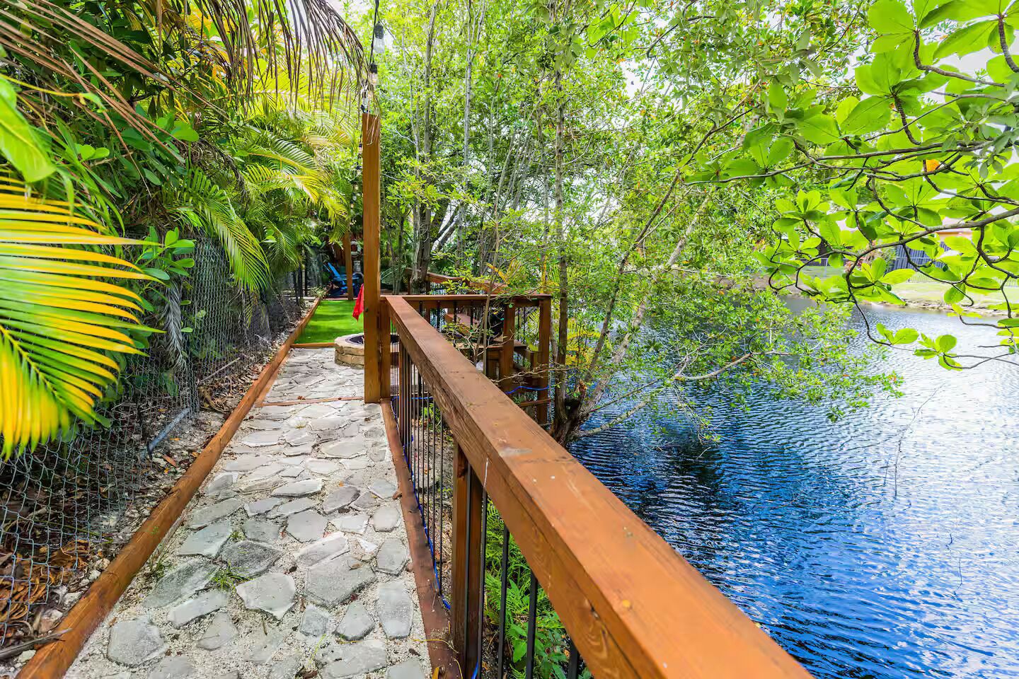 A wooden pathway leading to a water body, surrounded by lush greenery and a chain-link fence.