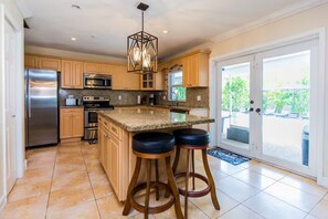 A modern kitchen with a central island, bar stools, stainless steel appliances, and a sliding glass door leading to an outdoor area. - Tropical 5BR Retreat ! Pool, Bar, Games & Nature (Sunrise)