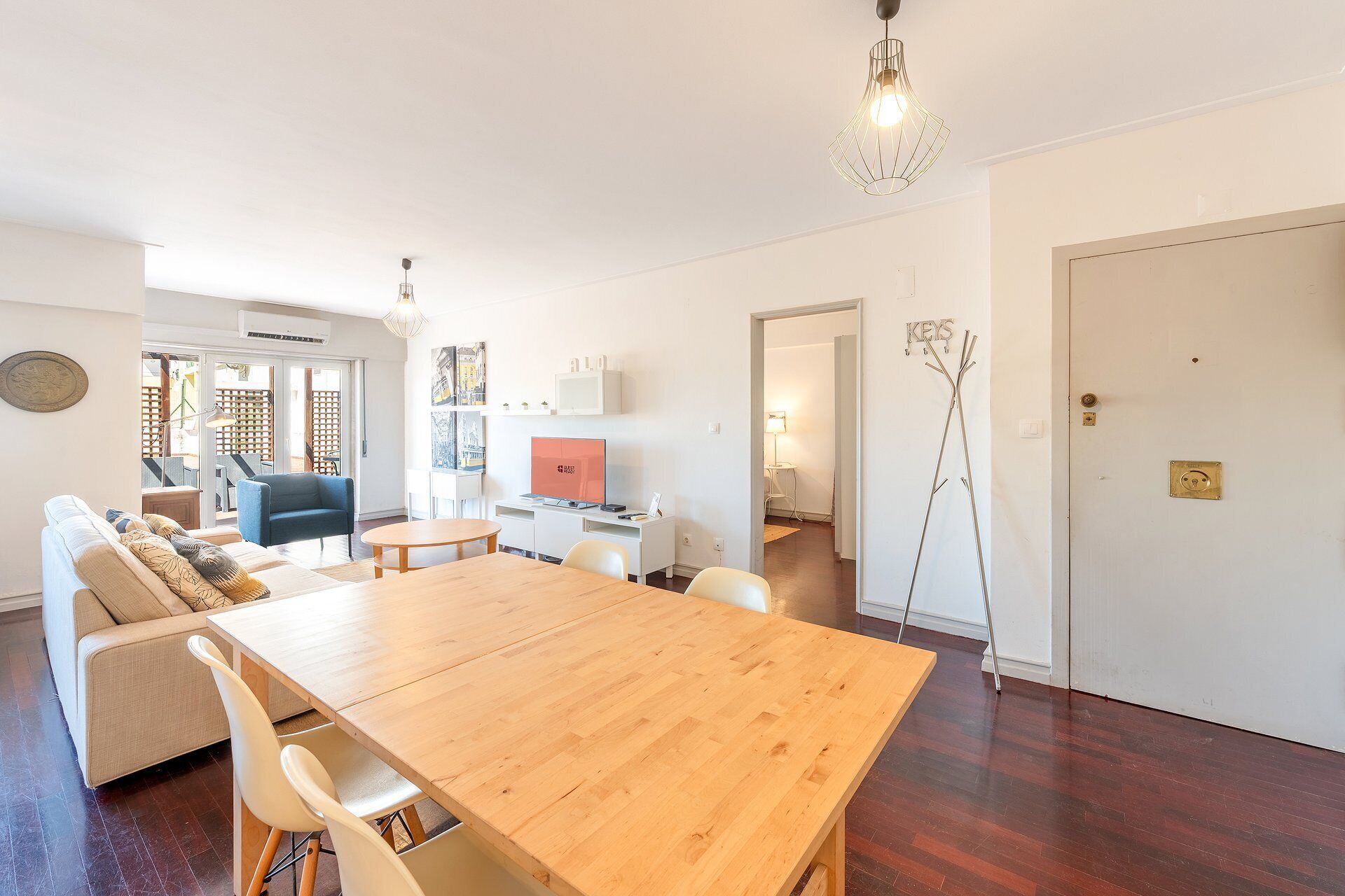 A modern living room with a wooden dining table, a sofa, and a blue armchair. There is a white door with a brass handle and a key holder on the wall.