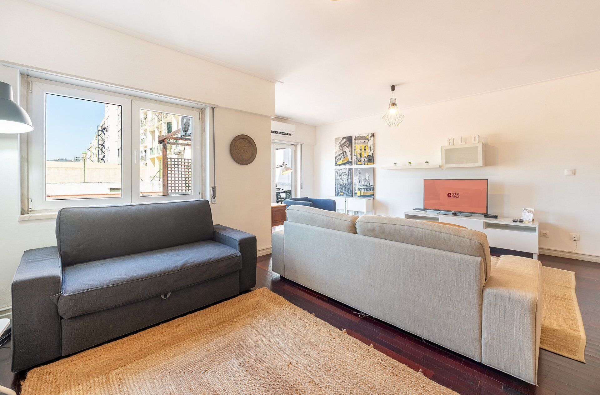 Un salon moderne avec un canapé gris, un canapé gris clair et un tapis marron. On y trouve un téléviseur posé sur un meuble blanc et une fenêtre donnant sur des immeubles.