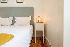 A neatly made bed with white linens and pillows, a bedside table with a spherical lamp, and a wooden floor. - GuestReady - Modern home near Rua Augusta (Lisbon)