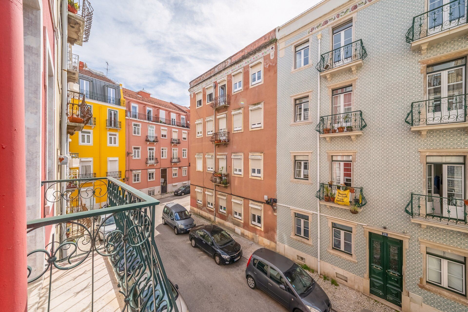 A narrow street with colorful buildings, parked cars, and balconies with plants.