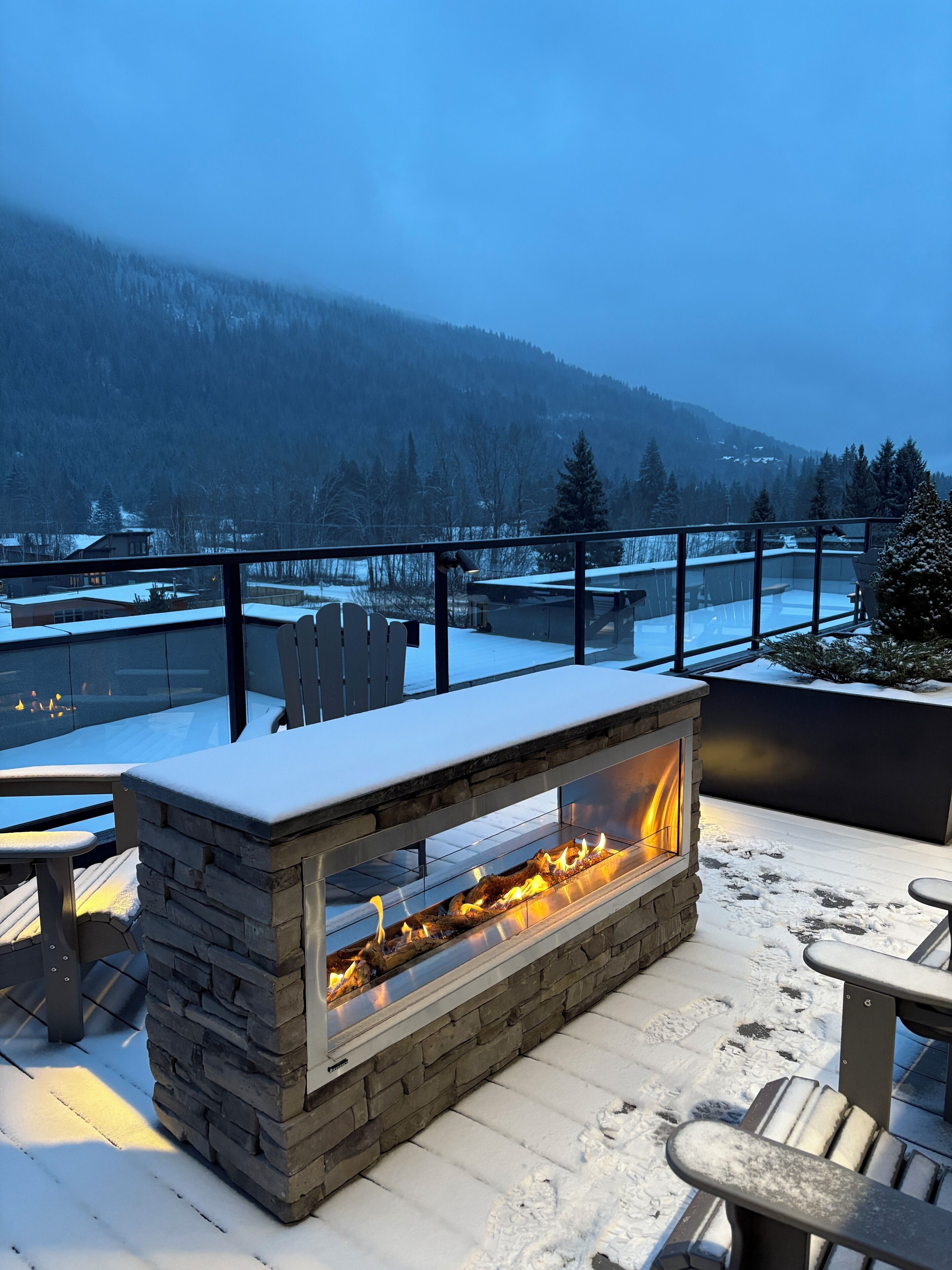 A stone fire feature with a glass enclosure, outdoor seating, and a view of snow-covered mountains.