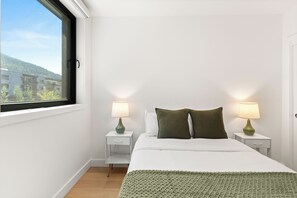 Un dormitorio con cama, dos mesitas de noche con lámparas, una ventana con vista a edificios y un plaid verde de punto sobre la cama.