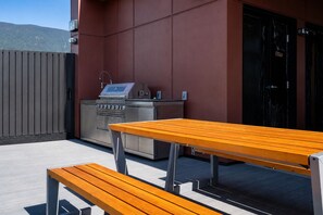 Een buitenpatio met een houten tafel en bank, een roestvrijstalen barbecue en een rode muur.
