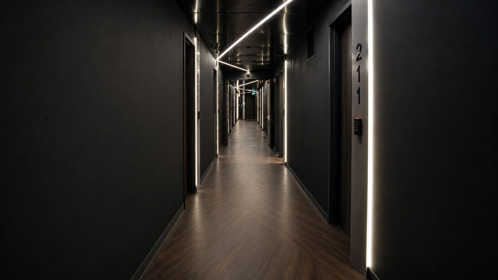 A modern hotel hallway with a dark floor, metallic walls, and illuminated ceiling lines.