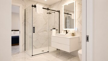 Un baño moderno con ducha envuelta en vidrio, tocador blanco con espejo y suelo de mármol.
