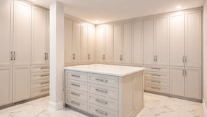 A modern kitchen with white cabinetry, a central island with drawers, and a marble floor. - Loft 1325 (Val-d'Or)