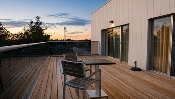 A wooden deck with a table and chairs, large windows, and a modern building facade.