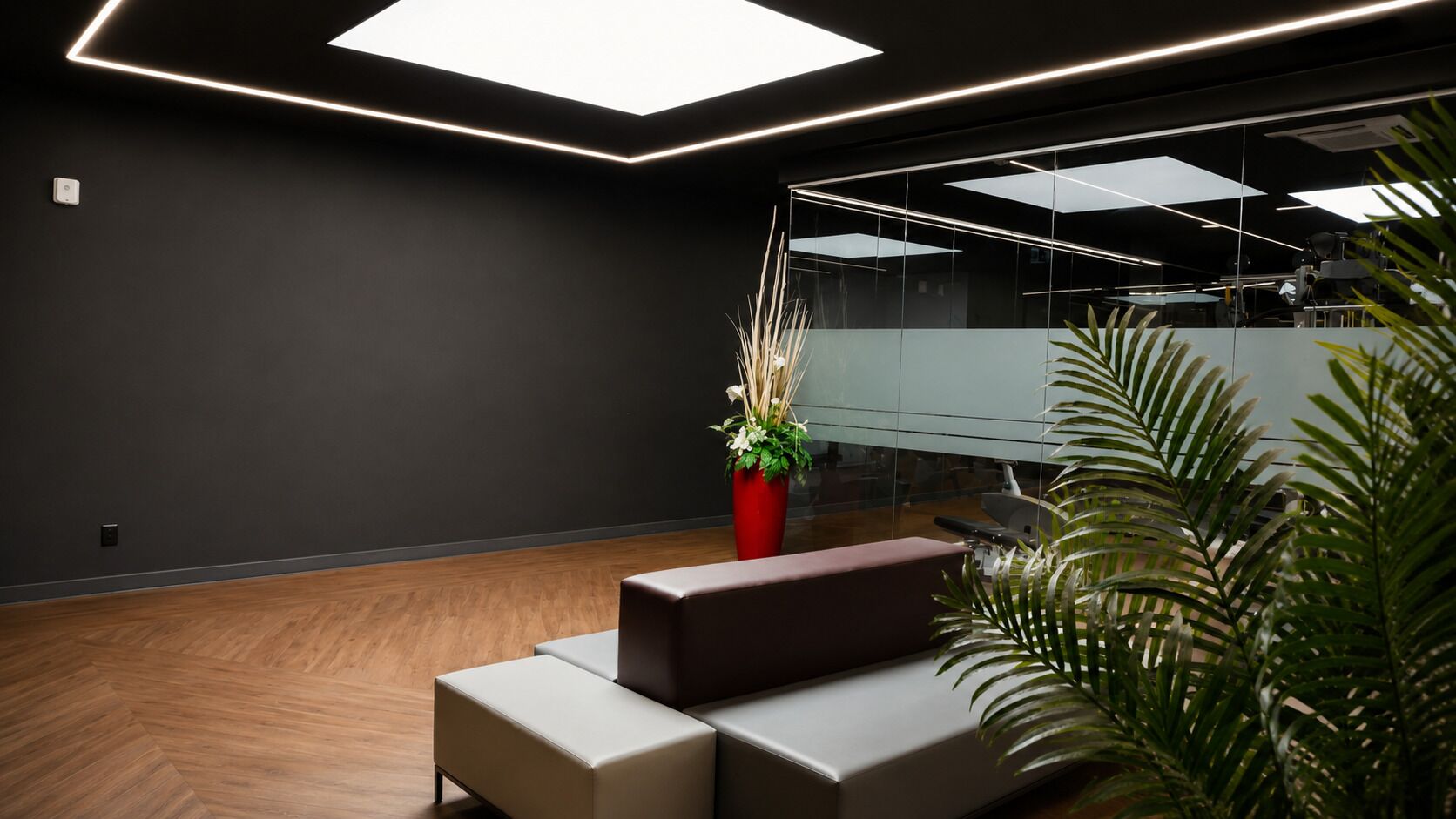 Um espaço de escritório moderno com piso de madeira em padrão herringbone, uma planta em vaso vermelho e uma área de estar com sofá marrom.