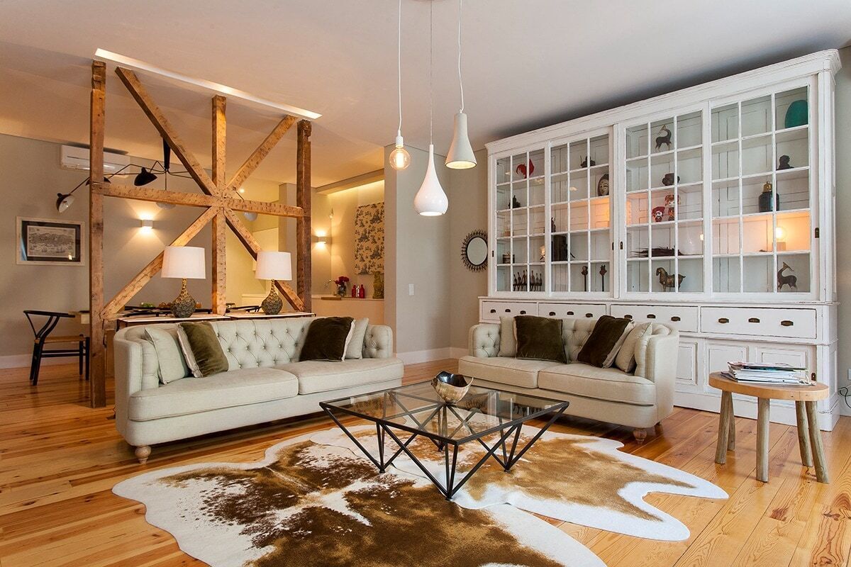 Ein Wohnzimmer mit zwei Sofas, einem Glastisch, einem Kuhfellteppich, einem weißen Schrank mit Glastüren und einer Holzbalkenkonstruktion.