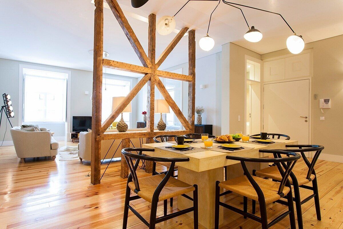 A dining area with a wooden table and chairs, a kitchen in the background, and a large window with natural light.