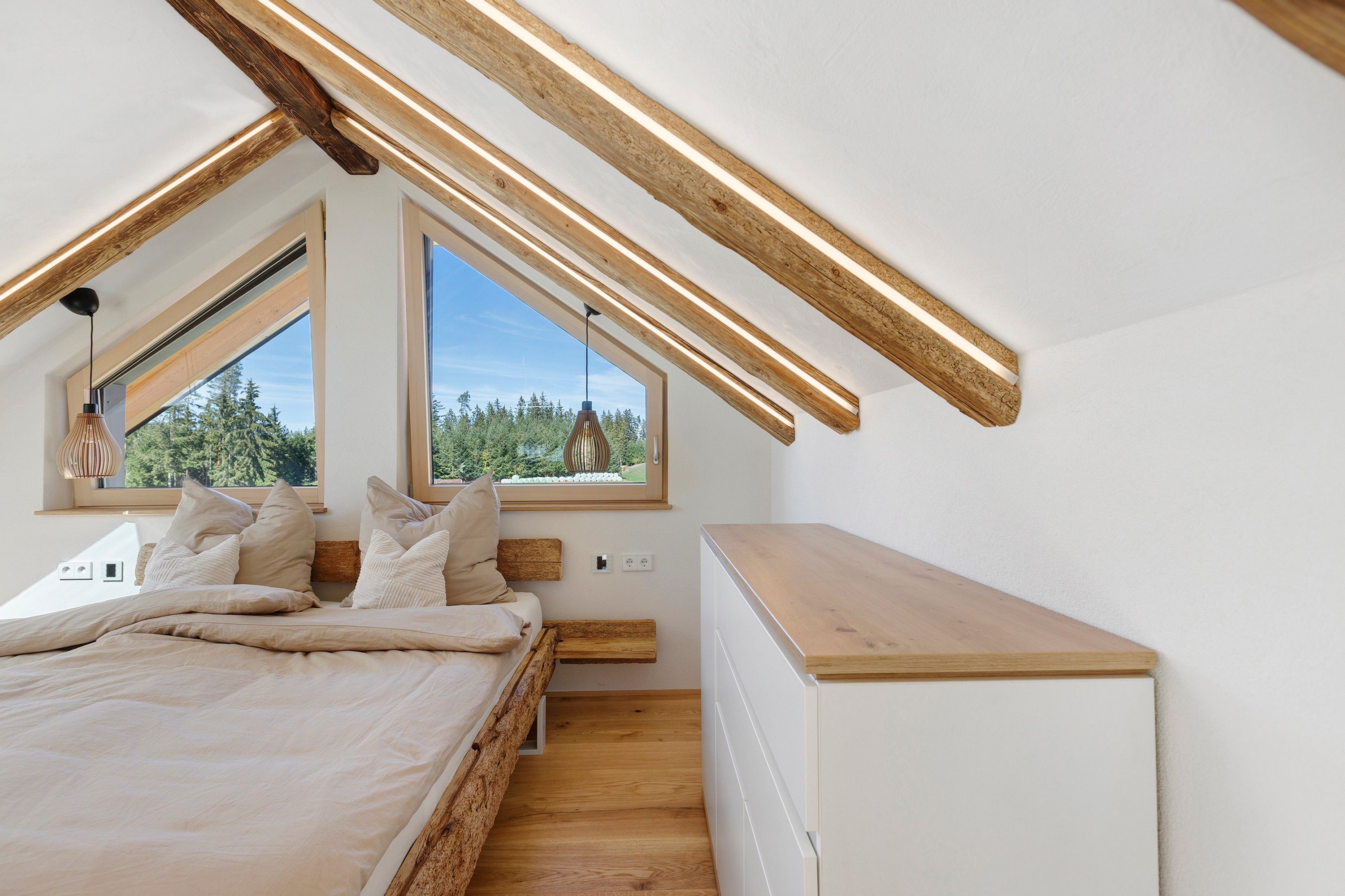 Une chambre avec un plafond en pente, des poutres en bois, un lit avec des oreillers, une table de chevet et une fenêtre donnant sur des arbres.