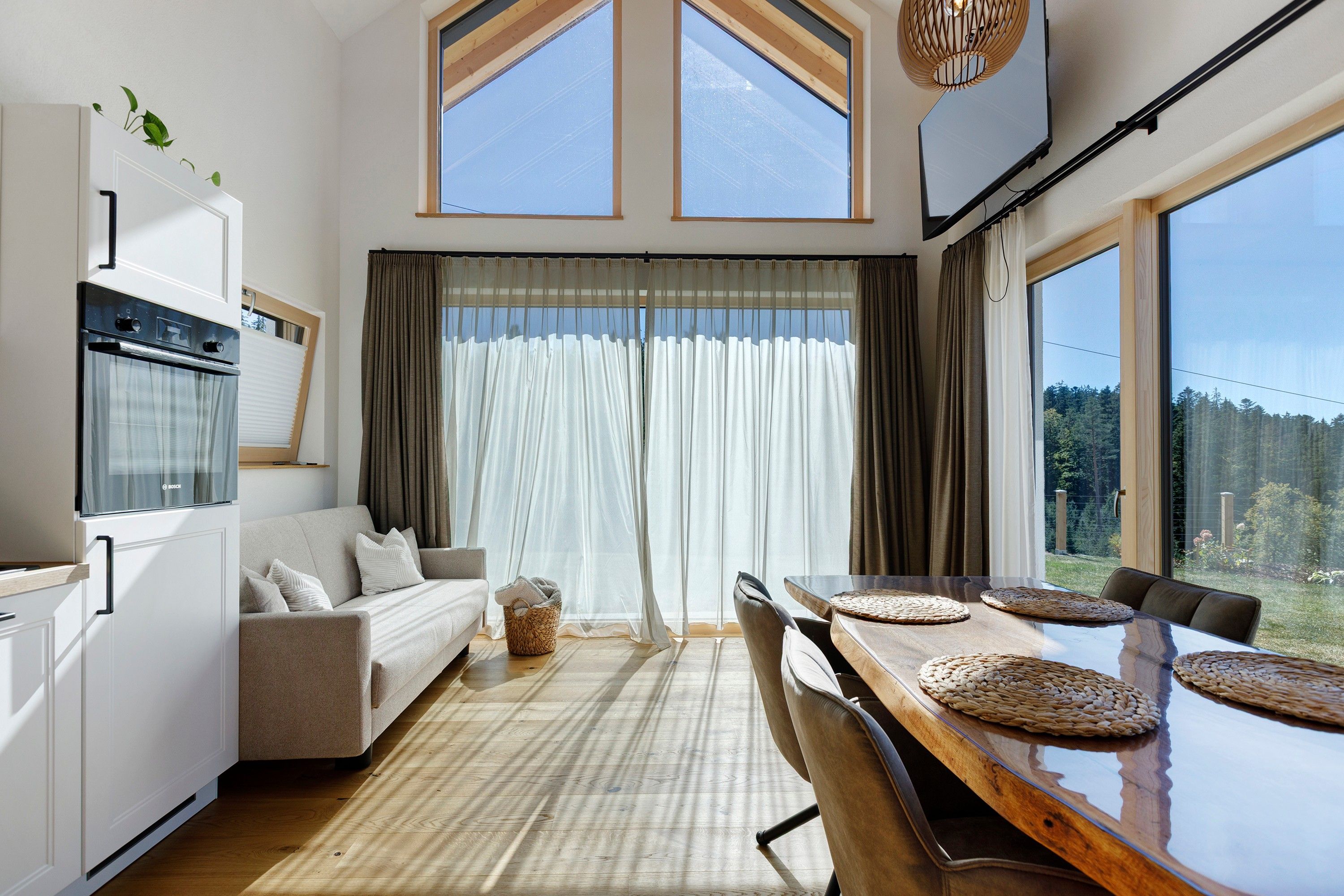 Eine moderne Küche mit Backofen, ein Essbereich mit Holztisch und gewebten Tischsets, eine Sitzecke und ein großes Fenster mit durchsichtigen Vorhängen.