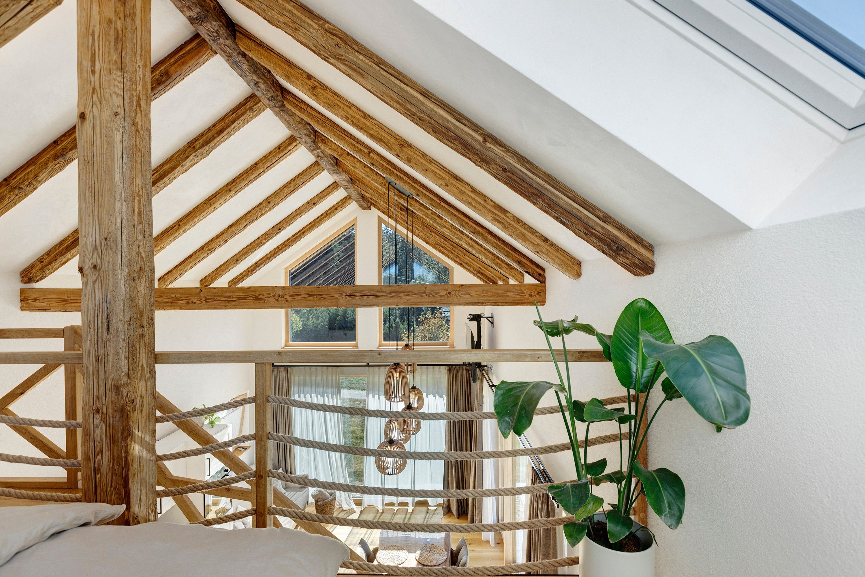 Ein Zimmer mit schräg verlaufender Holzbalkendecke, großen Fenstern und einer Seilvorrichtung.