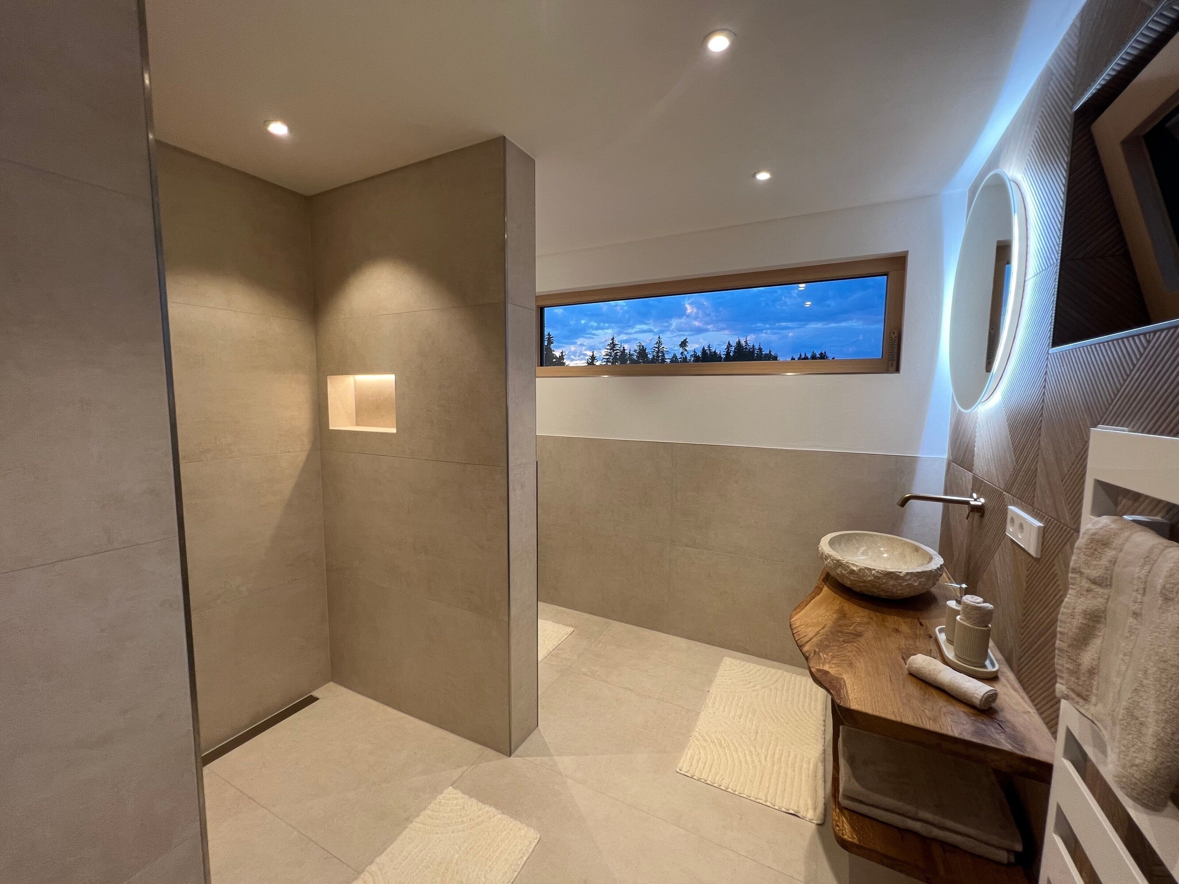 Ein modernes Badezimmer mit einem großen Spiegel, einem Holzkosmetikschrank und Blick auf den Himmel durch ein Fenster.