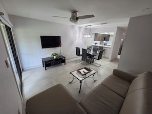 A modern living room with a flat-screen TV, a ceiling fan, a dining area with a glass table, and a kitchen in the background. - High Point World Resort  (Kissimmee)