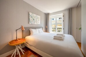 A hotel room with a large bed, two bedside lamps, a small wooden side table, a painting on the wall, and a view of the outside balcony. - GuestReady - Cosy stay near Money Museum (Lisbon)