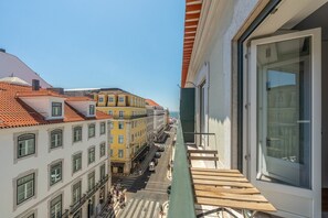 Een balkon met uitzicht op een straat vol gebouwen, een heldere lucht en een rustige zee in de verte.