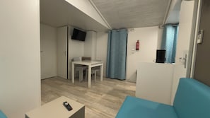A compact room with a bed, a small table, a chair, a TV mounted on the wall, and a window with curtains. - Sweet 6-Person Mobile Home – Le Neptune 4* Campground (Argelès-sur-Mer)