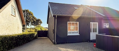 En boligejendom med en belagt indkørsel, et sort hus med en hvid dør, og et synligt brønddæksel.