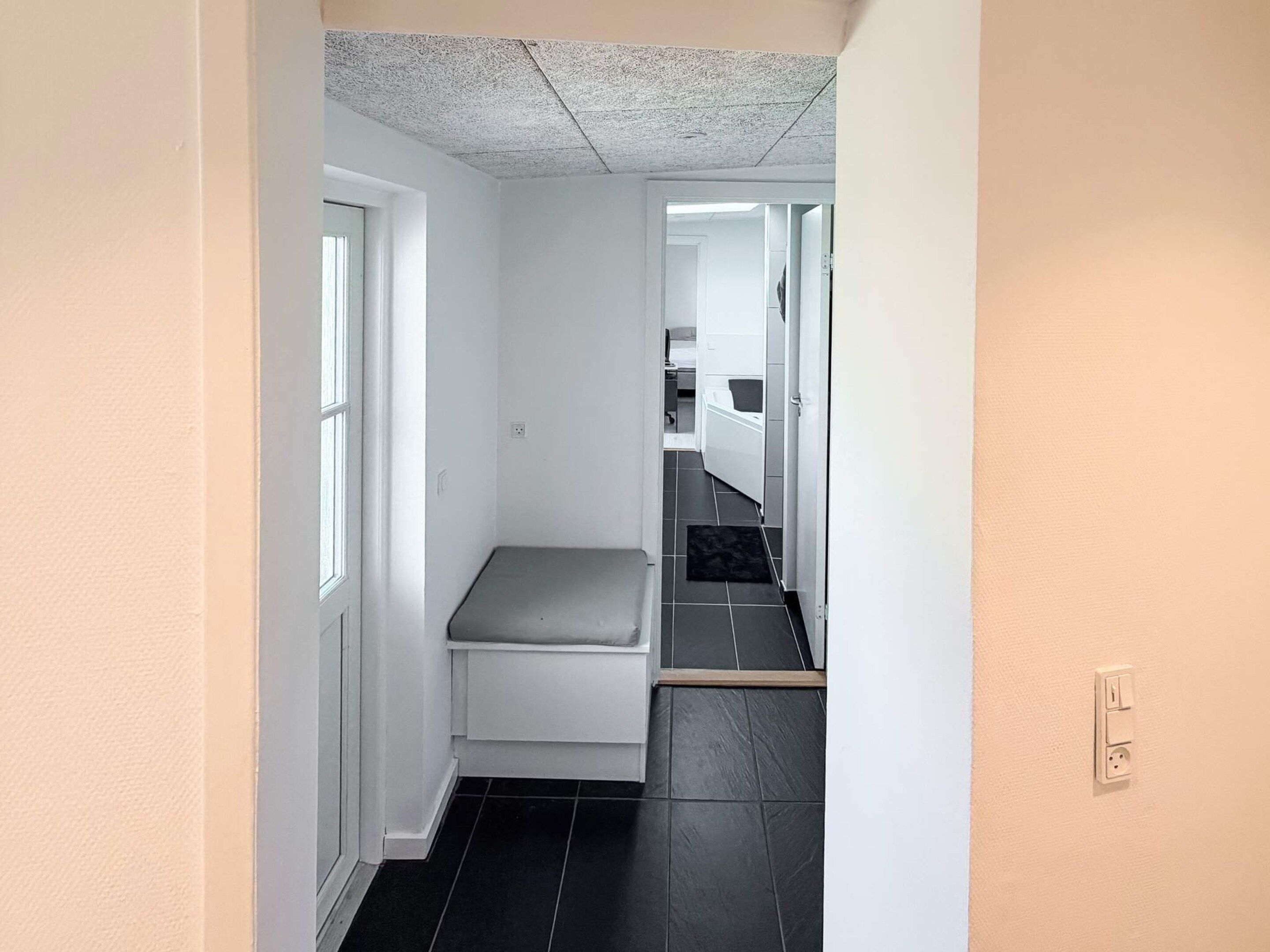A narrow hallway with a bench, a door leading to a bathroom, and a tiled floor.