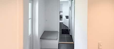 Um corredor estreito com um banco, uma porta de acesso a um banheiro e piso ladrilhado.