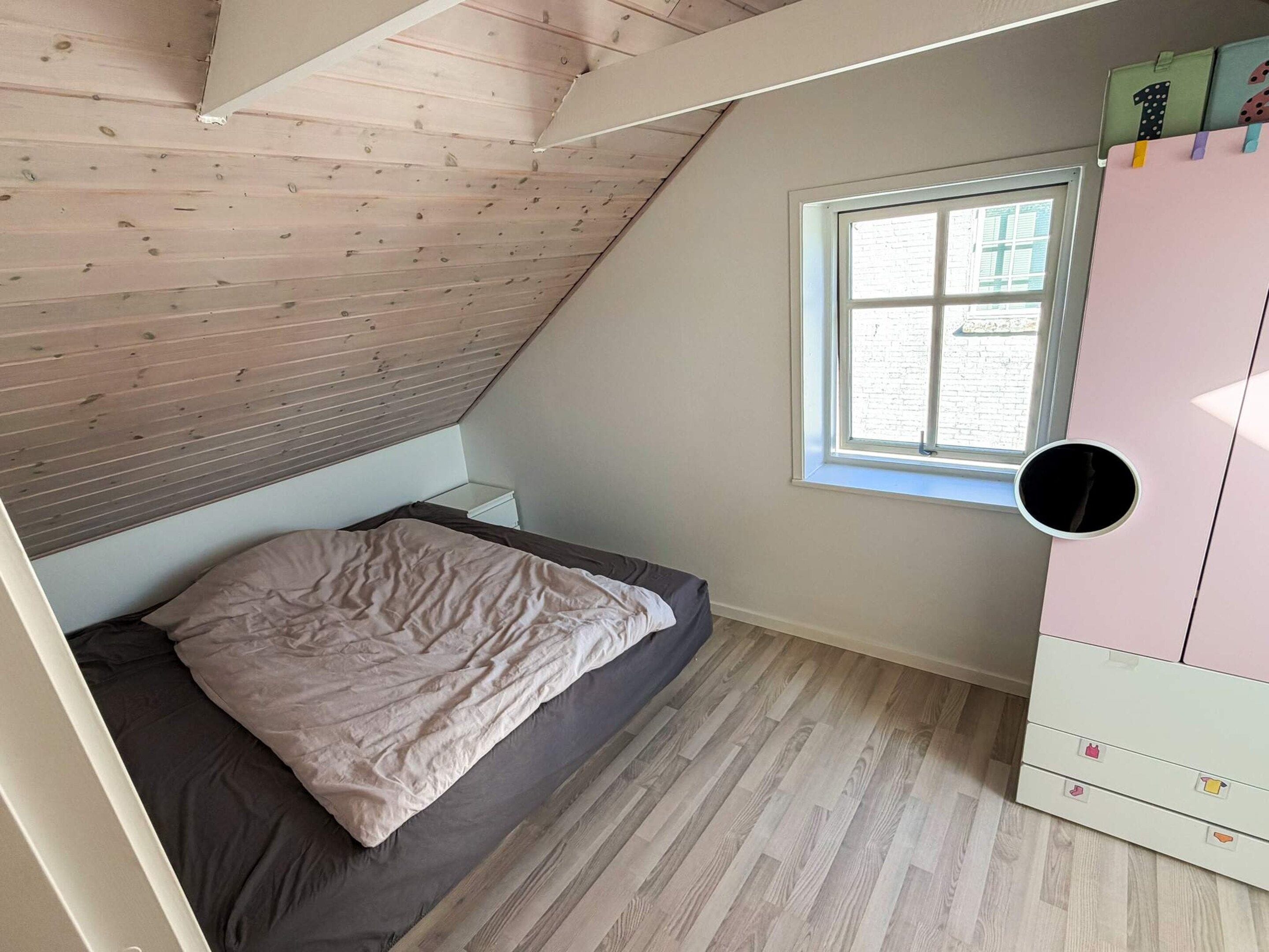 Une chambre avec un plafond en pente, un lit simple, une fenêtre avec des stores et une armoire dotée d’un miroir rond.