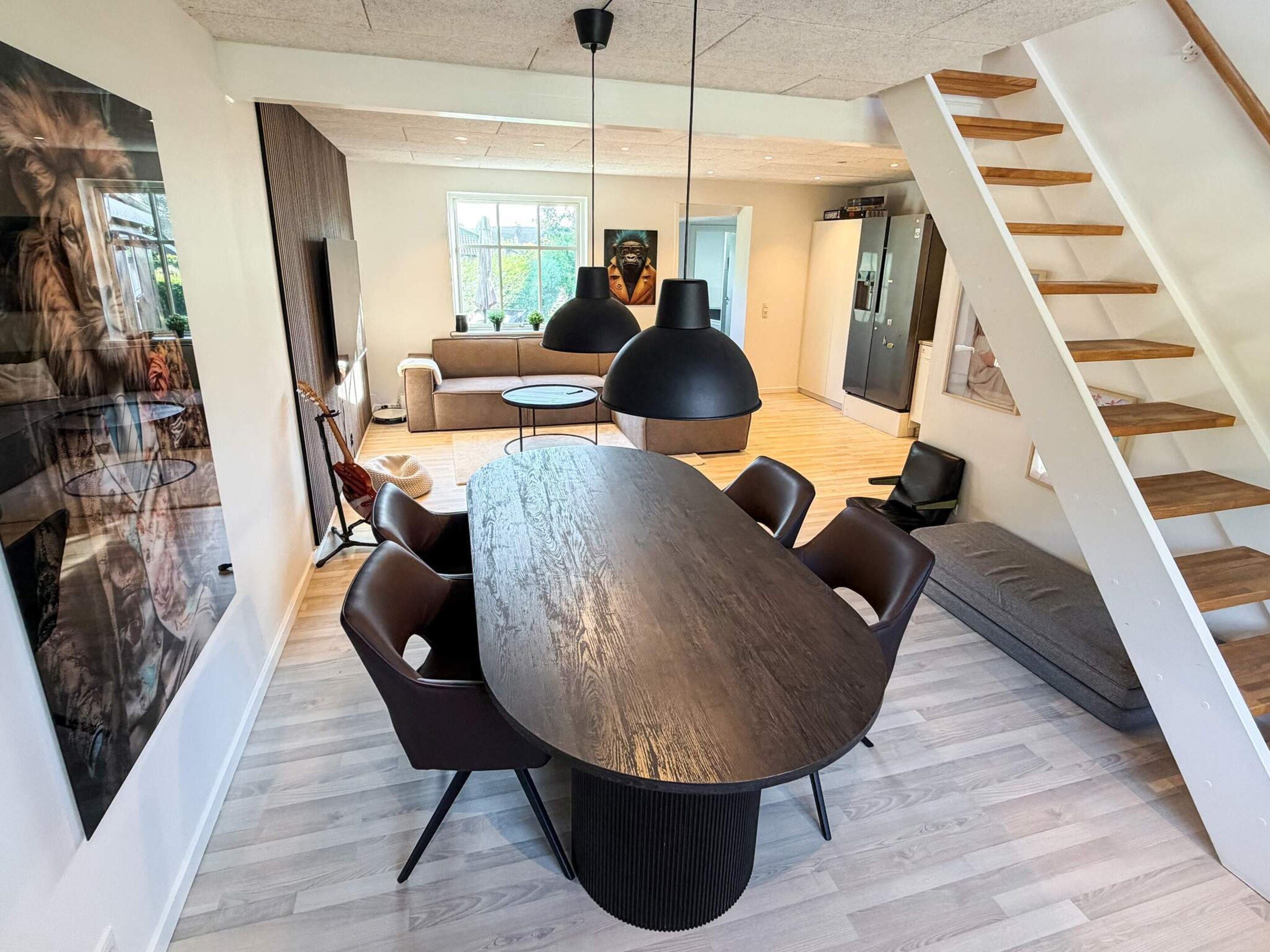 A modern living room with a large round table, dark chairs, a sofa, and a staircase.