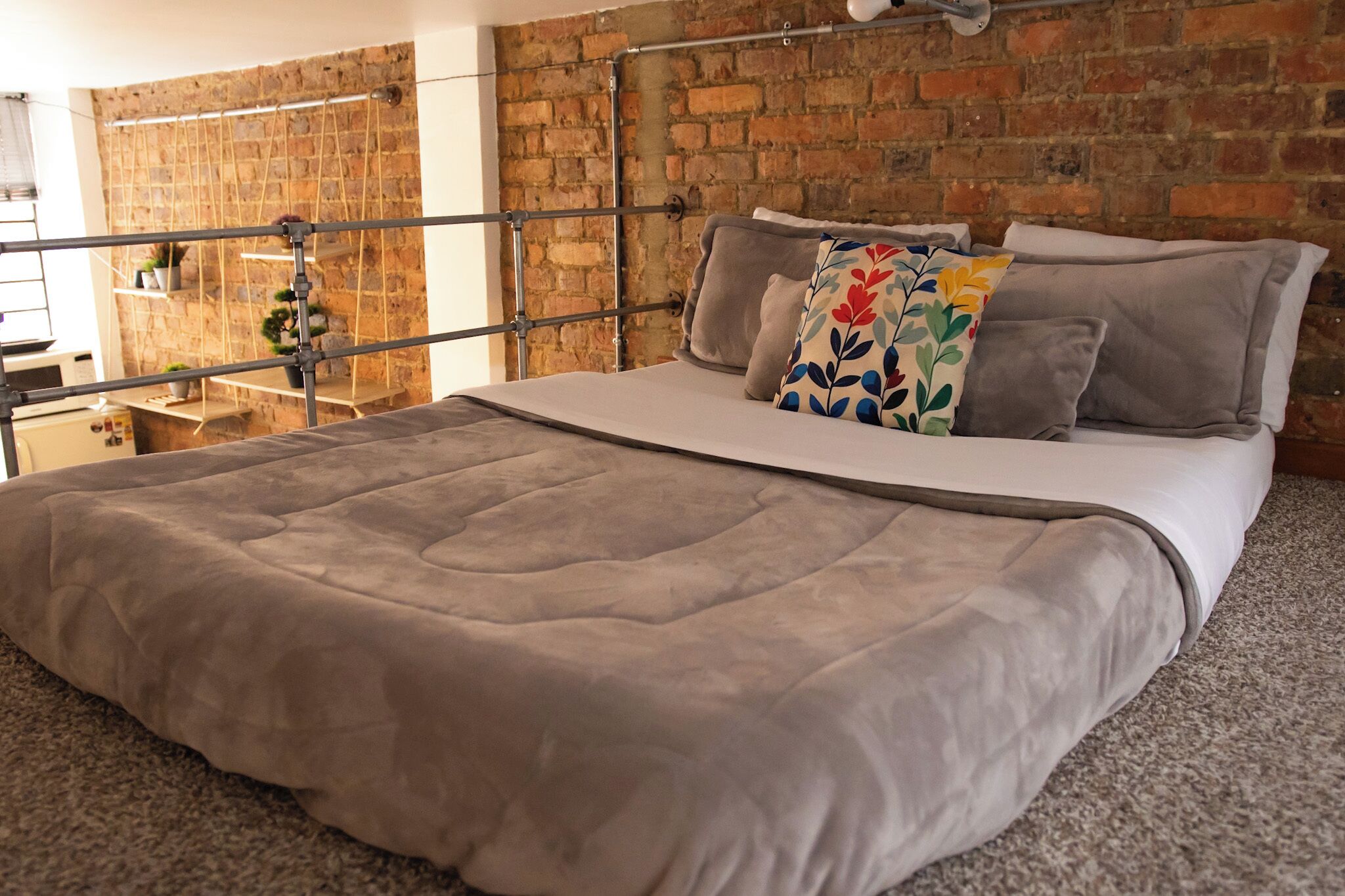 A bedroom with a brick wall, a metal headboard, a bed with a grey duvet, and a colorful pillow.