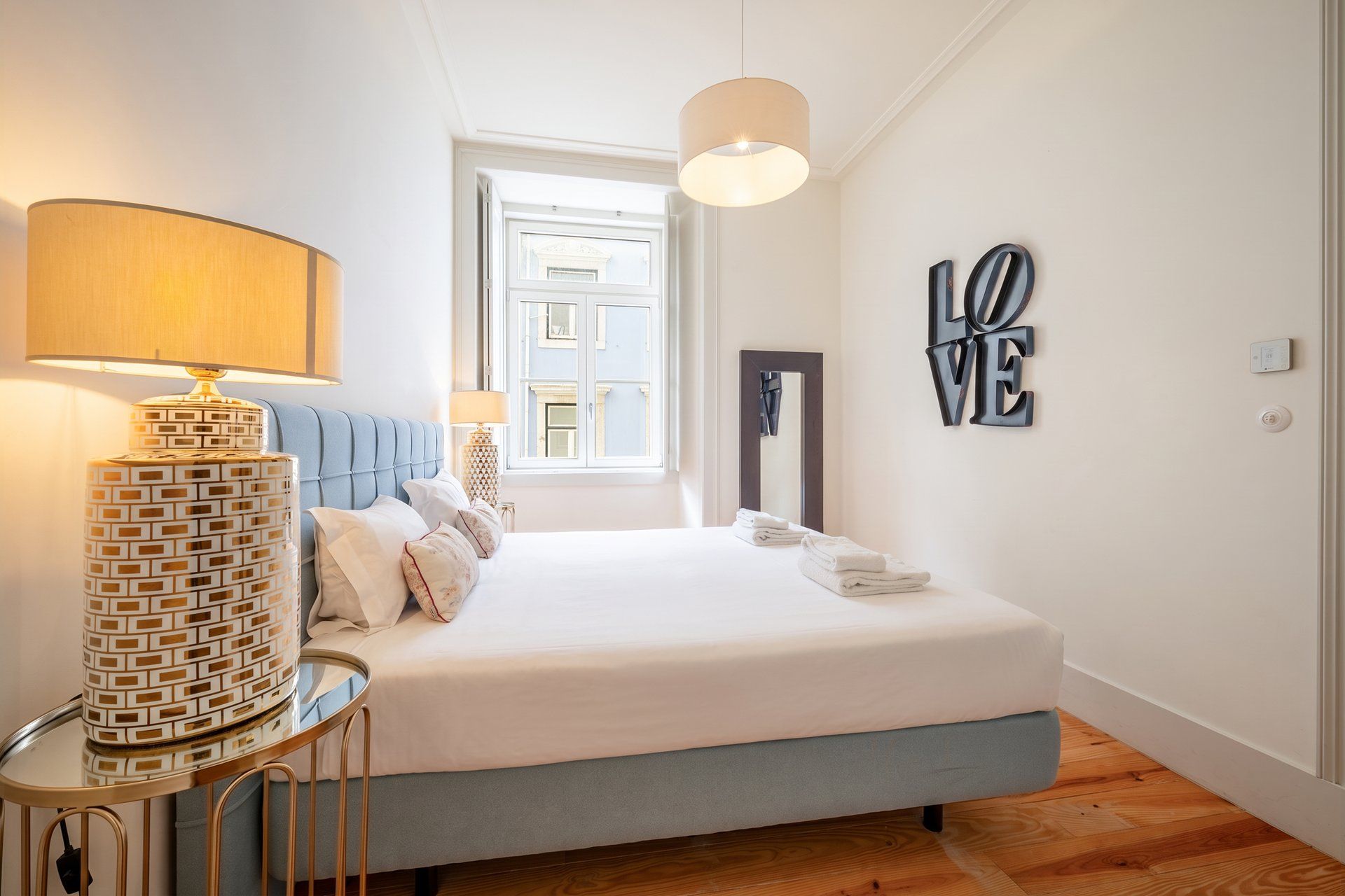 A neatly made bed with pillows, a bedside lamp, a wall-mounted mirror, a 'LOVE' sign, and a window with a view.
