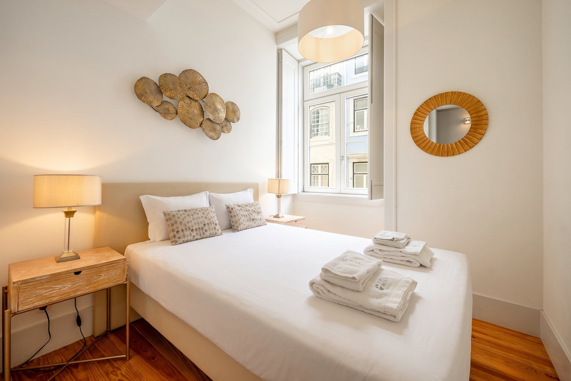 A hotel room with a bed, bedside lamps, a wall-mounted mirror, and a window with a view of buildings.