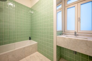 A bathroom with green tiles, a bathtub, a sink with a window above, and a showerhead. - GuestReady - Striking apt near Santa Justa Lift (Lisbon)