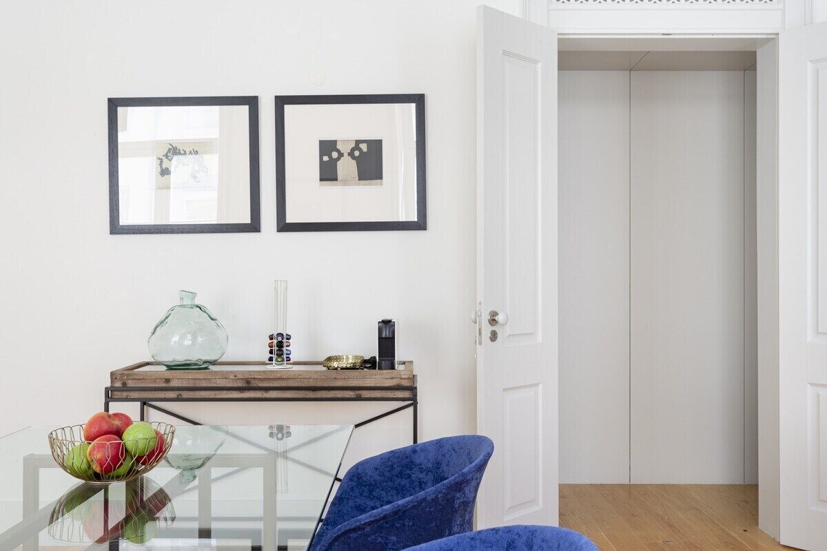 A room with a glass table, a blue chair, a wooden console table with decorative items, and two framed pictures on the wall.