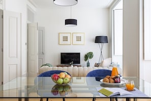 Ein modernes Wohnzimmer mit einem Glastisch, einer Couch, einem Fernseher und einem Essbereich mit einem Teller Essen und einem Glas Orangensaft.