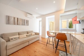 A modern living room with a sofa, two chairs, a coffee table, and a kitchen area with a window. - GuestReady - Sleek home near the Botanical Garden (Lisbon)