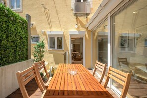 A wooden table with chairs in a sunlit outdoor area, a potted plant, and a building with a textured wall and windows. - GuestReady - Sleek home near the Botanical Garden (Lisbon)