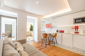 A modern kitchen with white cabinets, a microwave, and a red toaster. There is a dining area with a table and chairs, and a sofa in the foreground. - GuestReady - Sleek home near the Botanical Garden (Lisbon)