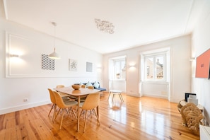 A spacious room with a wooden dining table, chairs, and a small round table. There are two windows with curtains, a ceiling light fixture, and wall-mounted artwork. - GuestReady - Trendy stay with sea view in Lapa (Lisbon)