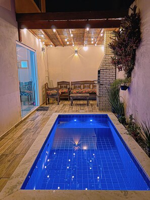 Una piscina con borde de azulejos, bancos de madera y techo con vigas y luces a la vista.