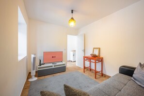 Ein modernes Wohnzimmer mit Fernseher, einem Standventilator, einem kleinen Tisch mit Korb und einem grauen Sofa.