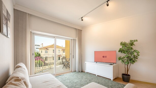 Una sala moderna con un sofá, un televisor sobre un mueble blanco, una planta en maceta y vistas de edificios a través de una puerta de cristal.