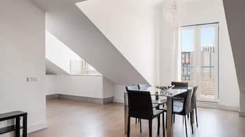 Zona pranzo moderna con tavolo e sedie in vetro, lampadario a sospensione e vista sulla città attraverso un'ampia finestra.