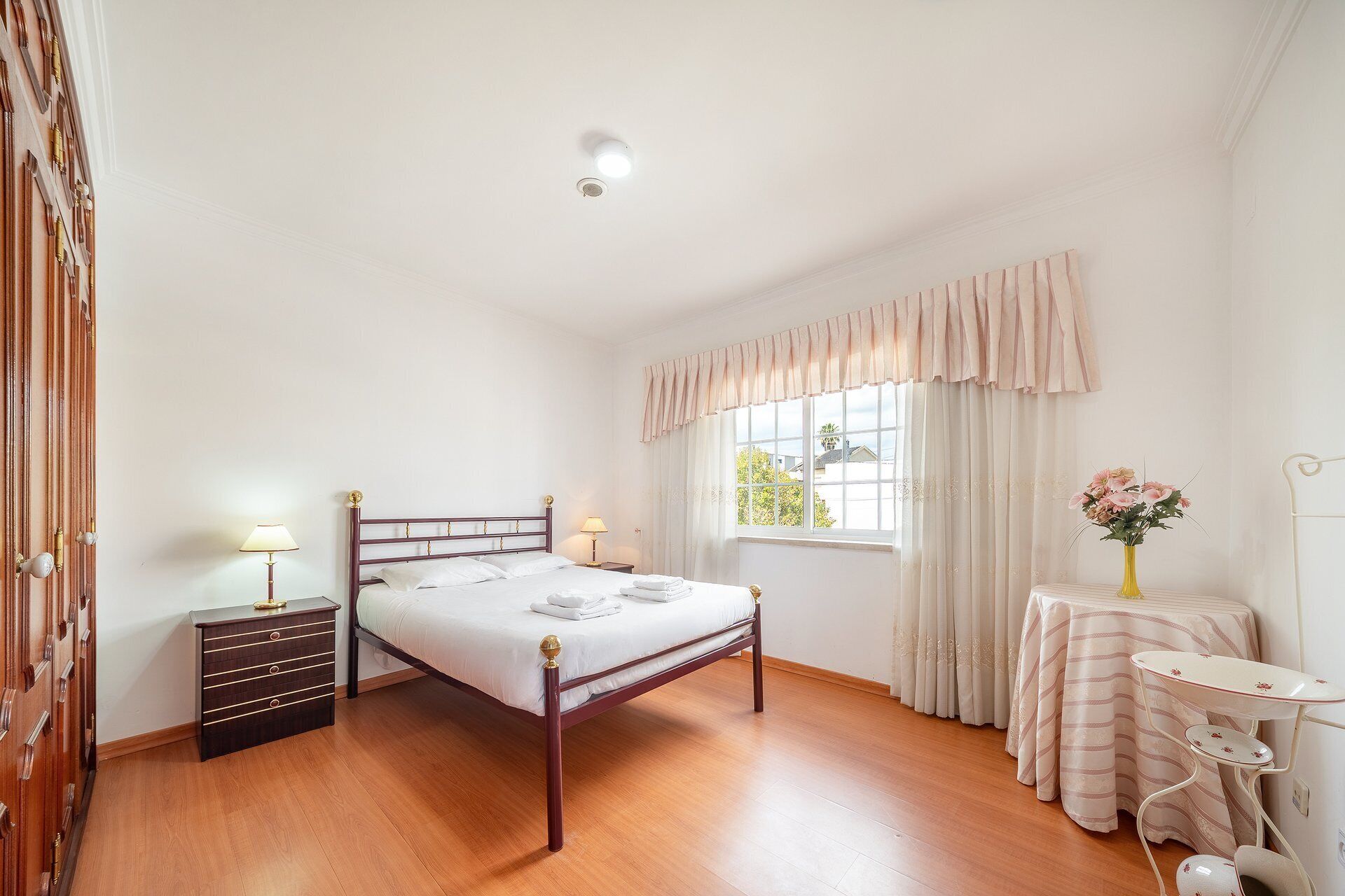 Un dormitorio con una cama de madera, una mesita de noche, una lámpara, una ventana con cortinas y una pequeña mesa con un florero.