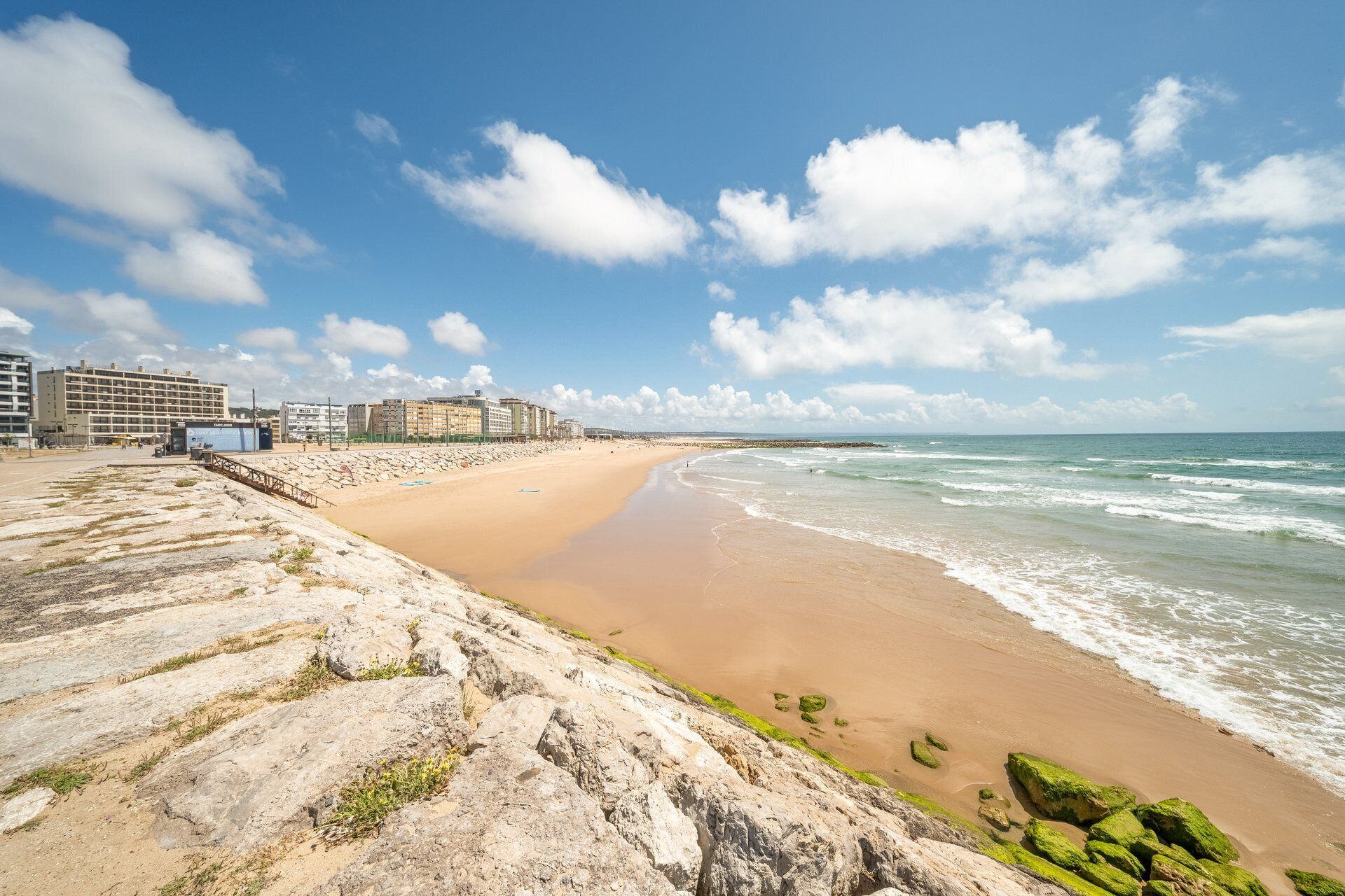 Una playa con costa rocosa, una playa de arena y una fila de edificios al fondo.
