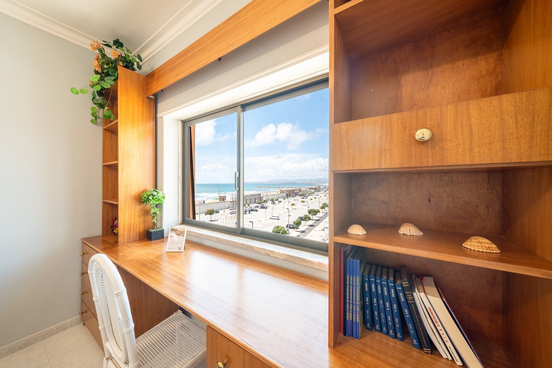 A room with a wooden desk, a chair, a window with a view of the beach, and shelves with books and decorative items.