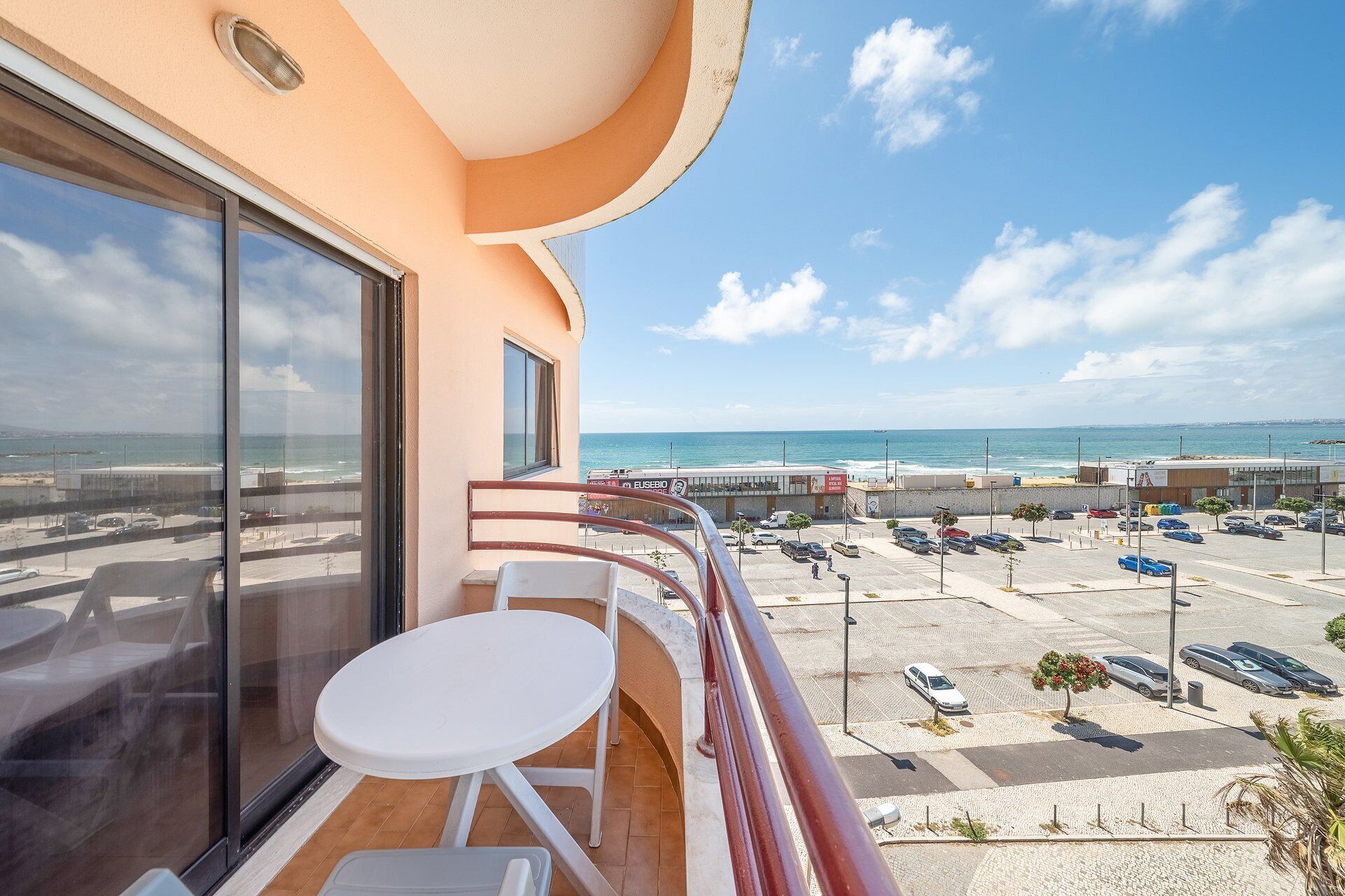 A balcony with a view of the sea, a parking lot, and a building with a sign.