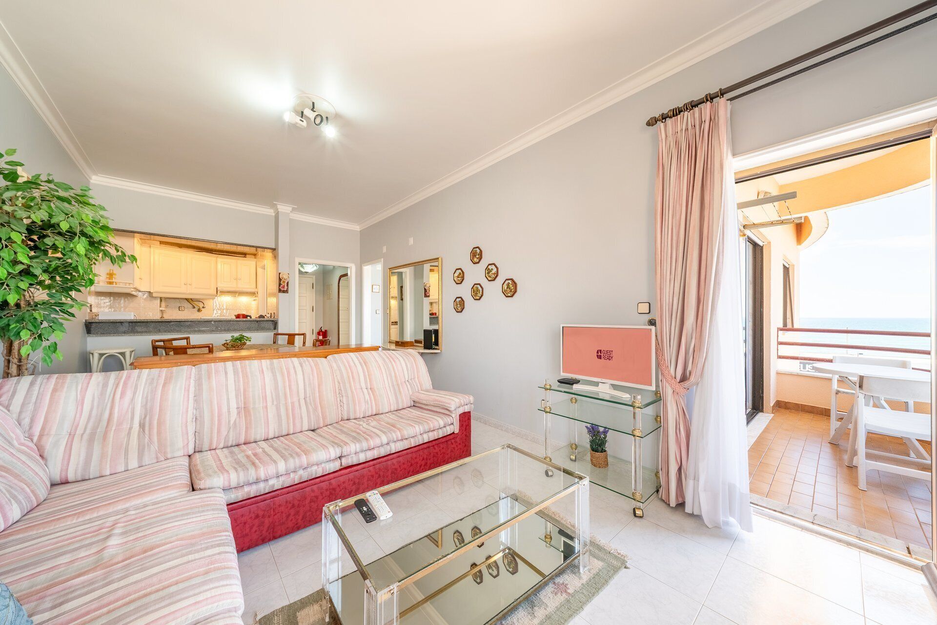 Un salon avec un canapé, une table basse en verre, un coin cuisine à l’arrière-plan et un balcon avec des rideaux.