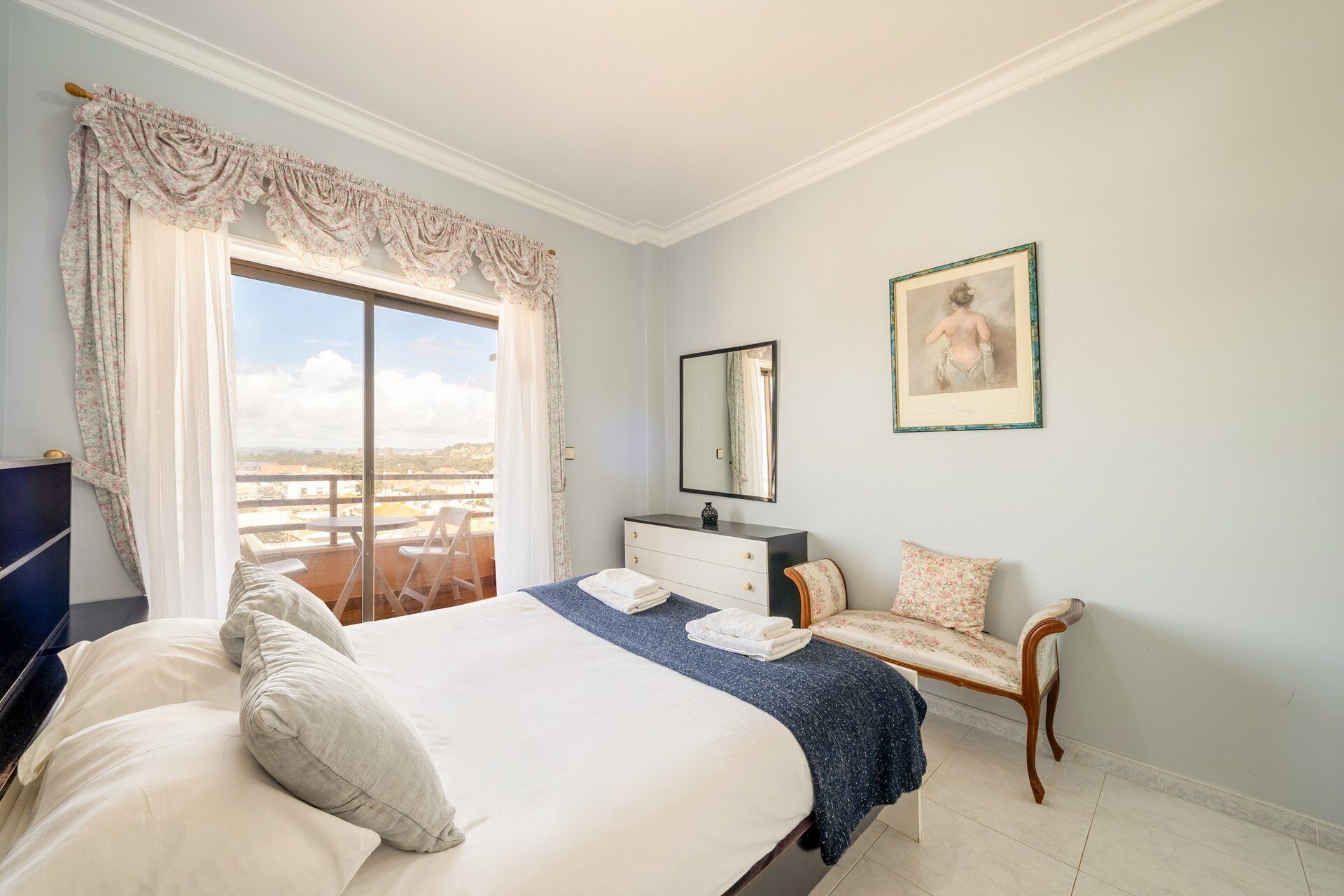 Un dormitorio con cama, una silla, cómoda, espejo y un cuadro en la pared.