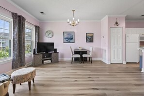 A living room with a television, a dining table with chairs, a chandelier, and a clock on the wall. - King Bed 1BR Condo | Golf View | Pool (Myrtle Beach)